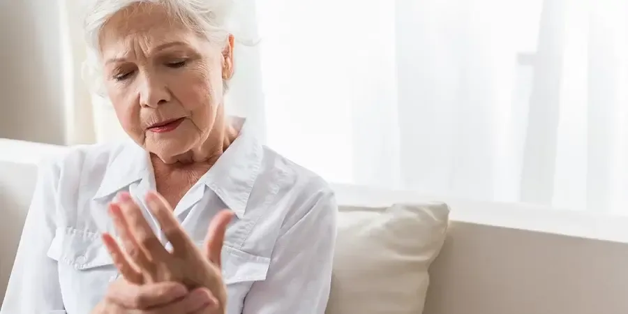 An elderly woman sits on a couch and holds her left hand with her right hand